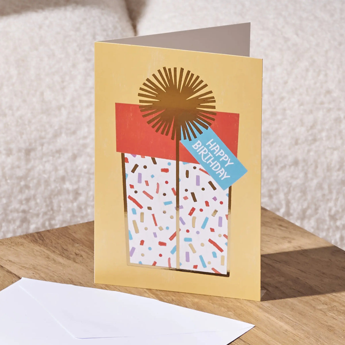 A gold birthday card standing open on a wooden surface, featuring a stylized gift box illustration with confetti and a blue 'Happy Birthday' tag. A white envelope lies in the foreground against a soft neutral background.