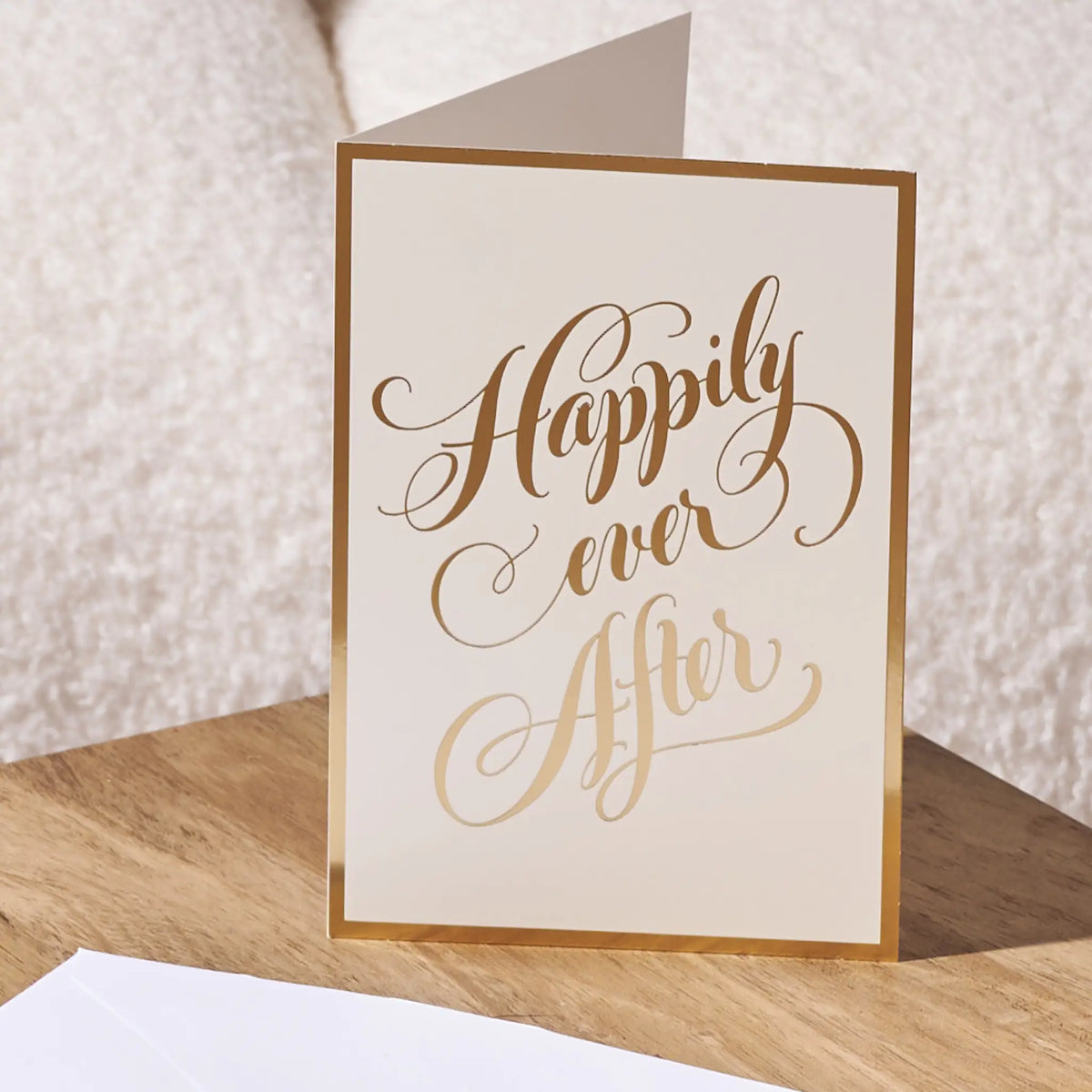 A cream-colored greeting card with elegant gold foil script reading 'Happily ever after' and a thin gold border, standing on a wooden surface against a soft beige textured background.