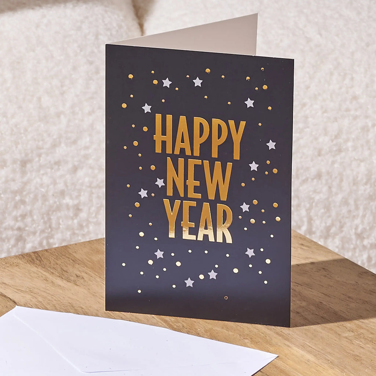 A folded greeting card standing upright on a wooden surface, with a white envelope in front. The card has a dark charcoal background with metallic gold and silver foil stars and the words 'HAPPY NEW YEAR' in large gold foil letters.