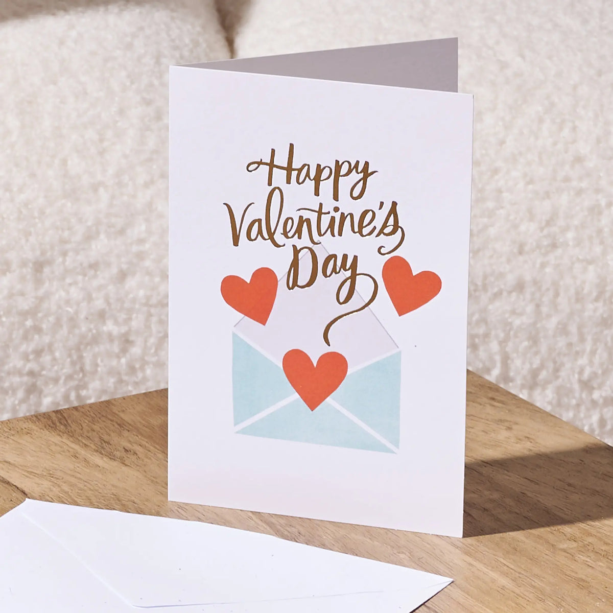 A white Valentine's Day greeting card standing on a wooden surface, printed with the words 'Happy Valentine's Day' above a light blue envelope and three red hearts. A matching white envelope lies in front, with a soft cream fabric background behind.