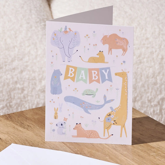 A baby greeting card with a whimsical safari animal illustration and a colorful 'BABY' banner, standing on a wooden surface with a white envelope in front.