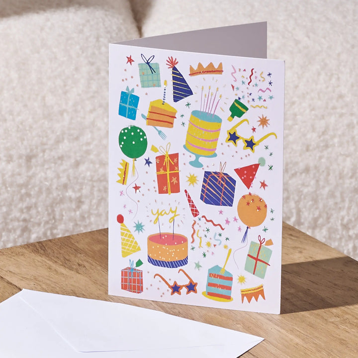 A white birthday card with colorful party-themed illustrations, standing on a wooden surface with a white envelope in front. The background shows a soft beige textured wall.