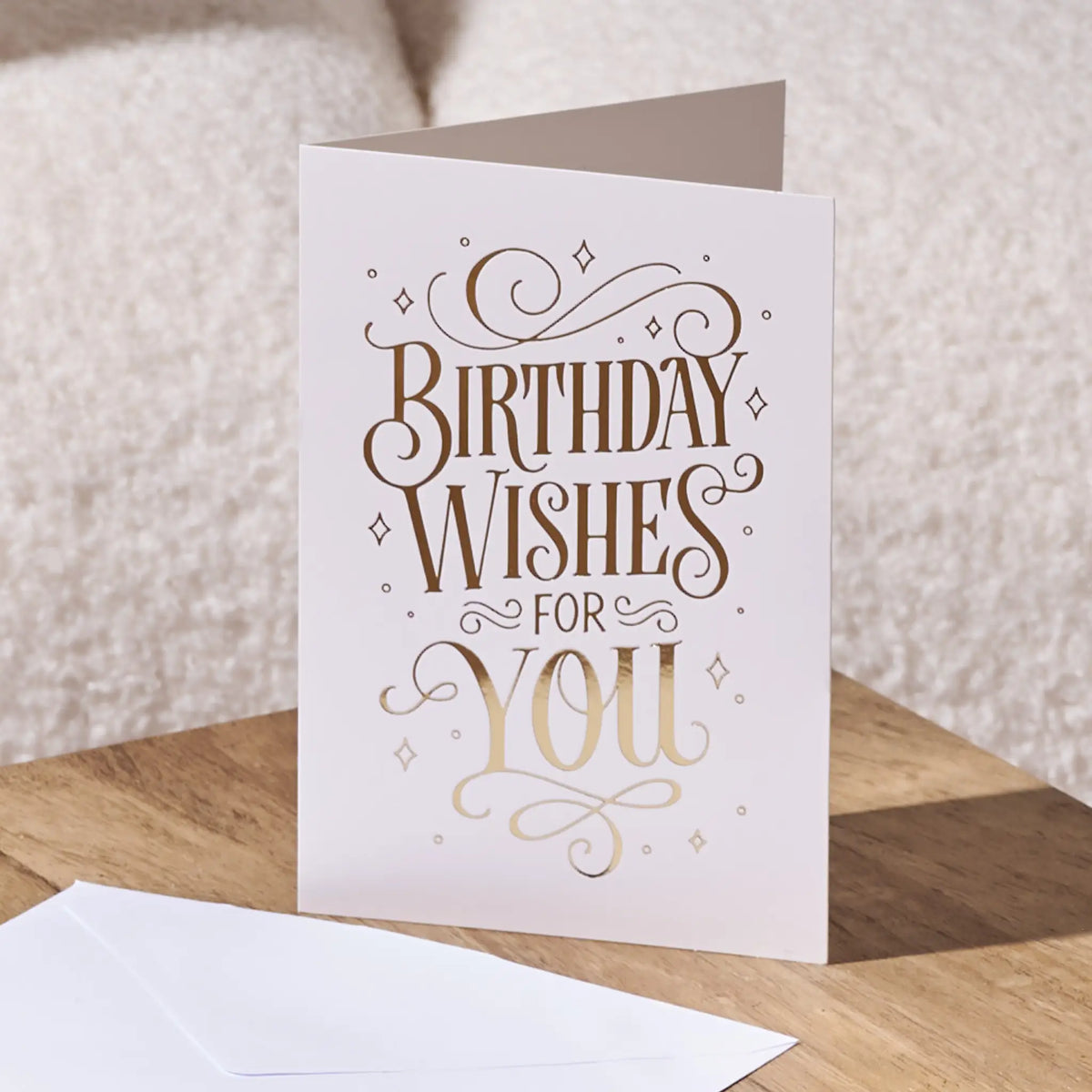 A white greeting card with elegant metallic gold foil lettering that reads 'BIRTHDAY WISHES FOR YOU', standing on a wooden surface with a white envelope in front and a soft beige textured background.