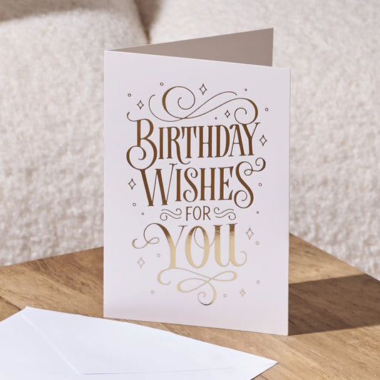 A white greeting card with elegant metallic gold foil lettering that reads 'BIRTHDAY WISHES FOR YOU', standing on a wooden surface with a white envelope in front and a soft beige textured background.