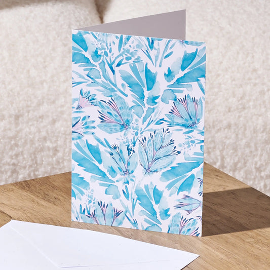 A standing greeting card with a white cover printed in a blue and light purple botanical pattern, shown on a wooden surface with a white envelope in front and a soft beige textured background.