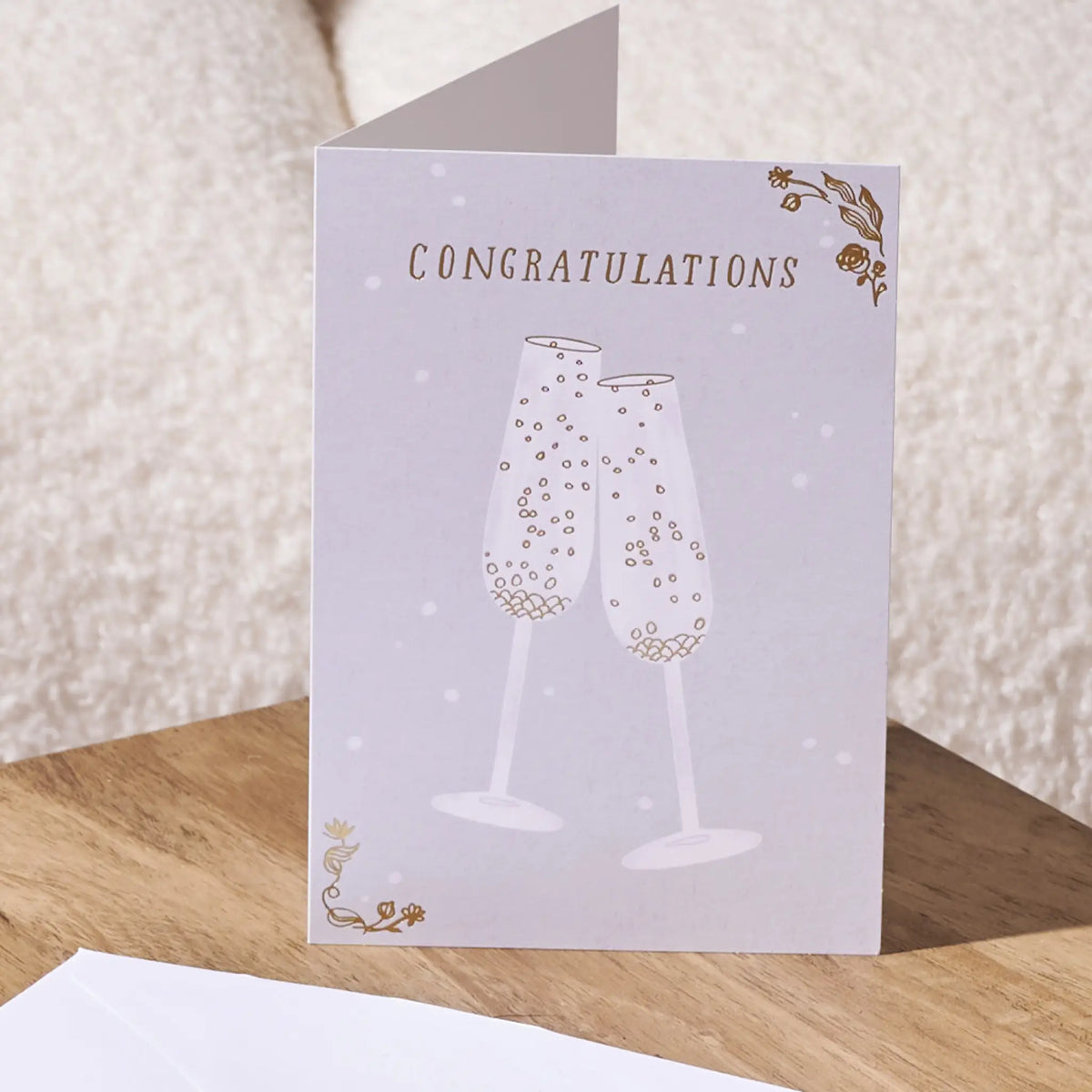 A folded congratulations card standing upright on a wooden surface, with a white envelope partly visible in the foreground. The card has a pale lavender background, gold floral corner accents, and an illustration of two champagne glasses with bubbles.