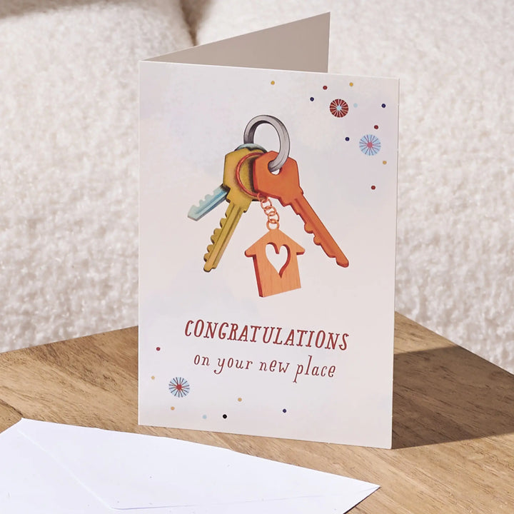 A folded congratulations card standing on a wooden surface, showing a set of keys with a house-shaped keychain and the text 'CONGRATULATIONS on your new place.' An envelope is placed in front, with a soft neutral background.