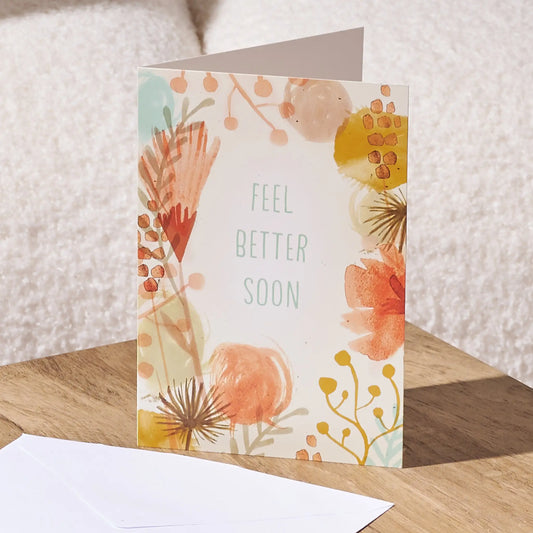 A folded greeting card standing on a wooden surface, printed with the words 'FEEL BETTER SOON' in light teal lettering and surrounded by soft watercolor floral and botanical illustrations in peach, orange, yellow, and muted blue. A white envelope lies in front, with a soft neutral background behind.