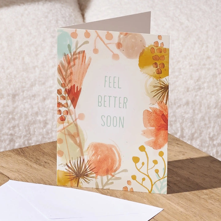 A folded greeting card standing on a wooden surface, printed with the words 'FEEL BETTER SOON' in light teal lettering and surrounded by soft watercolor floral and botanical illustrations in peach, orange, yellow, and muted blue. A white envelope lies in front, with a soft neutral background behind.