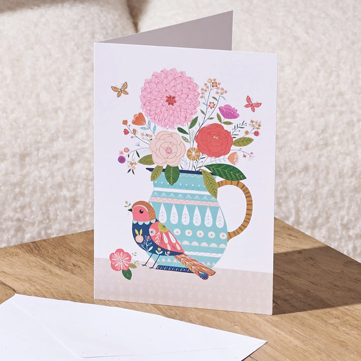 A folded greeting card standing open on a wooden surface, featuring a colorful illustration of a blue patterned pitcher filled with pink and red flowers, with a pink bird perched below. A white envelope lies in front, and a soft beige textured background is visible behind.