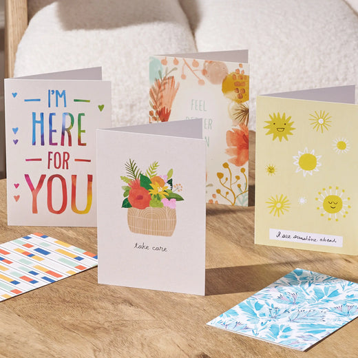 A set of greeting cards displayed on a wooden surface in front of a white textured sofa. The cards feature various colorful designs, including a rainbow 'I'm Here For You' card, a basket of flowers with 'Take care' text, a floral card, a yellow card with smiling suns, and patterned cards in the foreground.