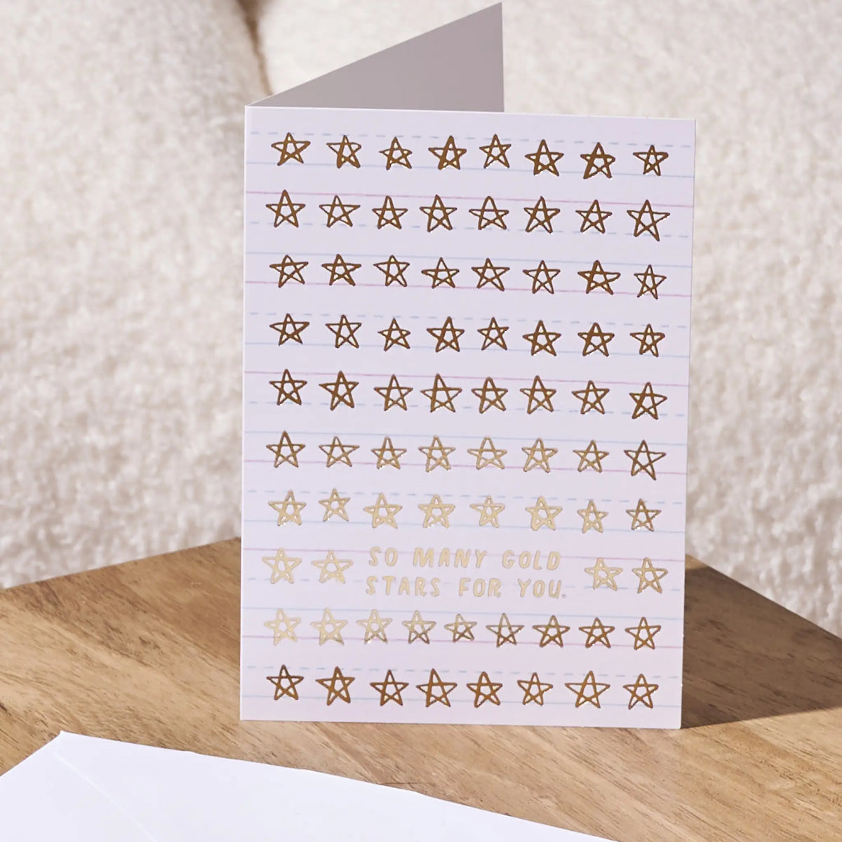 A lined greeting card with rows of gold star stickers and the phrase 'SO MANY GOLD STARS FOR YOU.' printed on the front, shown standing on a wooden surface with a neutral textured background.