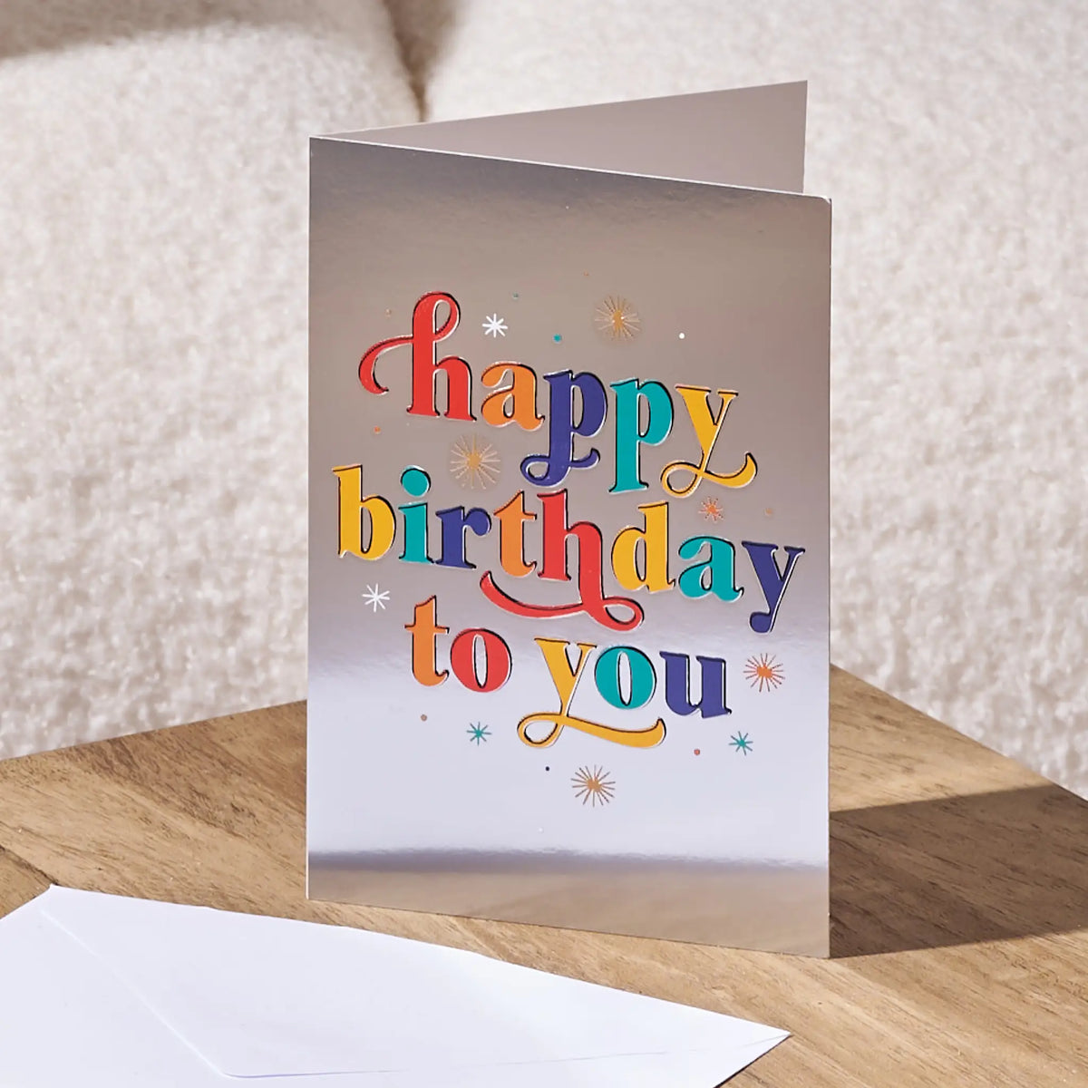 A folded greeting card standing on a wooden surface, with a white envelope in front. The card has a metallic silver background and colorful playful lettering that reads 'happy birthday to you' with small starburst accents.
