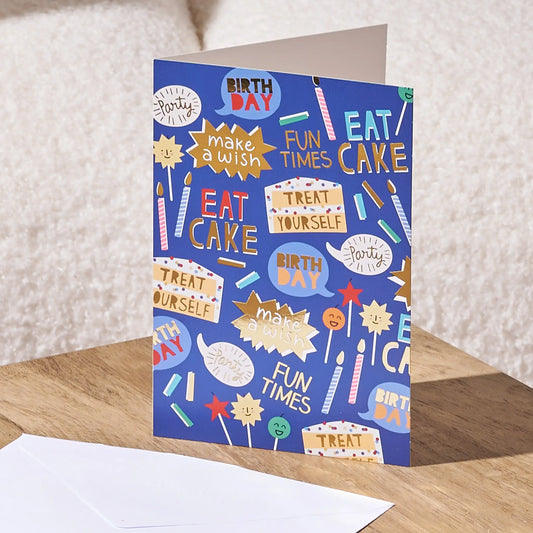 A blue birthday greeting card with a playful collage of party-themed illustrations and phrases like 'BIRTH DAY', 'EAT CAKE', and 'make a wish', shown standing on a wooden surface with a white envelope in front.