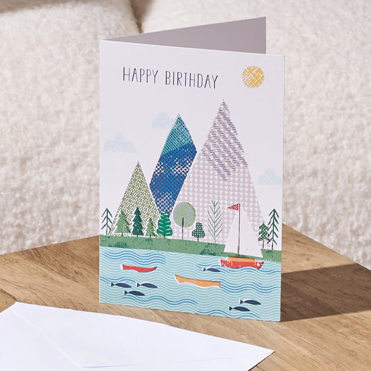 A folded birthday card standing on a wooden surface, showing a colorful illustrated landscape with mountains, trees, boats, fish, and the words “HAPPY BIRTHDAY.” An envelope lies in front, with a soft cream fabric background behind it.