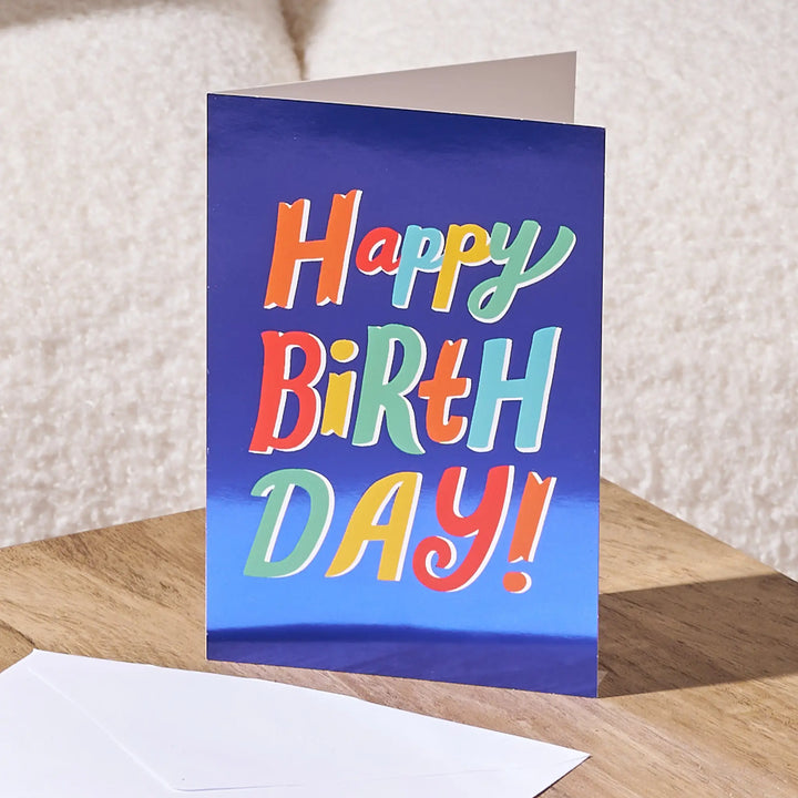 A colorful 'Happy Birthday!' greeting card standing upright on a wooden surface, with a white envelope in front and a soft beige textured background behind it.