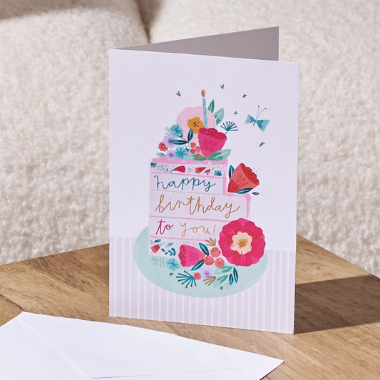 A folded birthday card standing on a wooden surface, featuring a watercolor-style illustration of a decorated cake with flowers and the words 'happy birthday to you!'. A white envelope lies in the foreground, with a soft beige textured background behind it.
