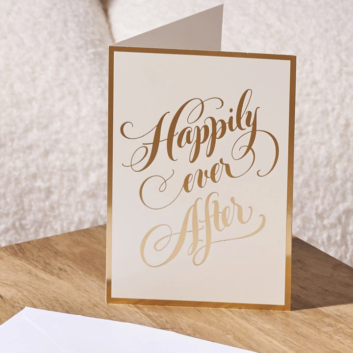 A cream-colored greeting card with elegant gold foil script reading 'Happily ever after' and a thin gold border, standing on a wooden surface against a soft beige textured background.