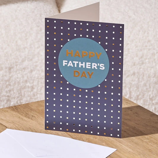 A folded Father's Day greeting card standing on a wooden surface, with a white envelope in front. The card has a dark navy background with a pattern of small white and gold dots, and a large light blue circle in the center reading 'HAPPY FATHER'S DAY' in gold and white lettering.