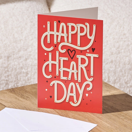 A red greeting card with 'HAPPY HEART DAY' in large white decorative lettering, standing on a wooden surface with a white envelope in front and a soft neutral background.