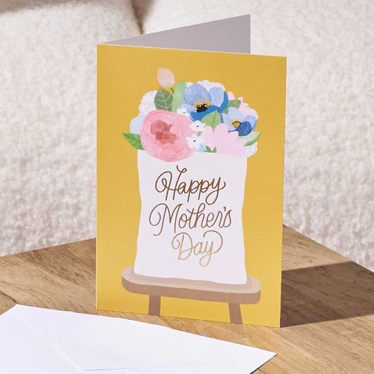 A yellow Mother's Day greeting card standing on a wooden surface, featuring a white sack filled with pink, blue, and white flowers and the words 'Happy Mother's Day' in metallic gold script. A white envelope is placed in front, with a soft beige textured background behind.