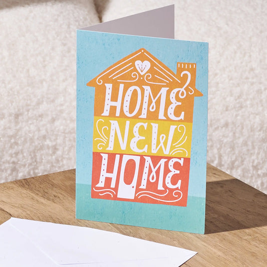 A folded greeting card standing upright on a wooden surface, with a white envelope in front. The card has a light blue background and features a house-shaped graphic with the words 'HOME NEW HOME' in white lettering over orange, yellow, and coral-red sections.