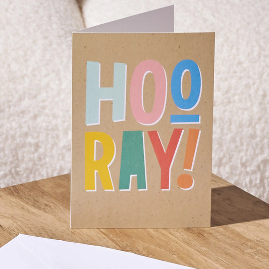 A folded kraft paper greeting card standing upright on a wooden surface, printed with the word 'HOO RAY!' in large pastel letters. A white envelope is partially visible in the foreground against a soft beige textured background.