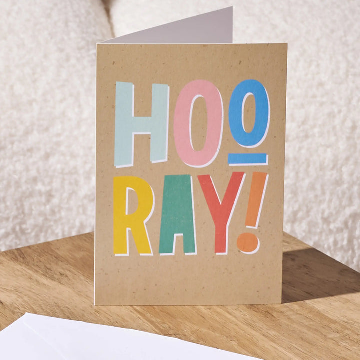 A folded kraft paper greeting card standing upright on a wooden surface, printed with the word 'HOO RAY!' in large pastel letters. A white envelope is partially visible in the foreground against a soft beige textured background.
