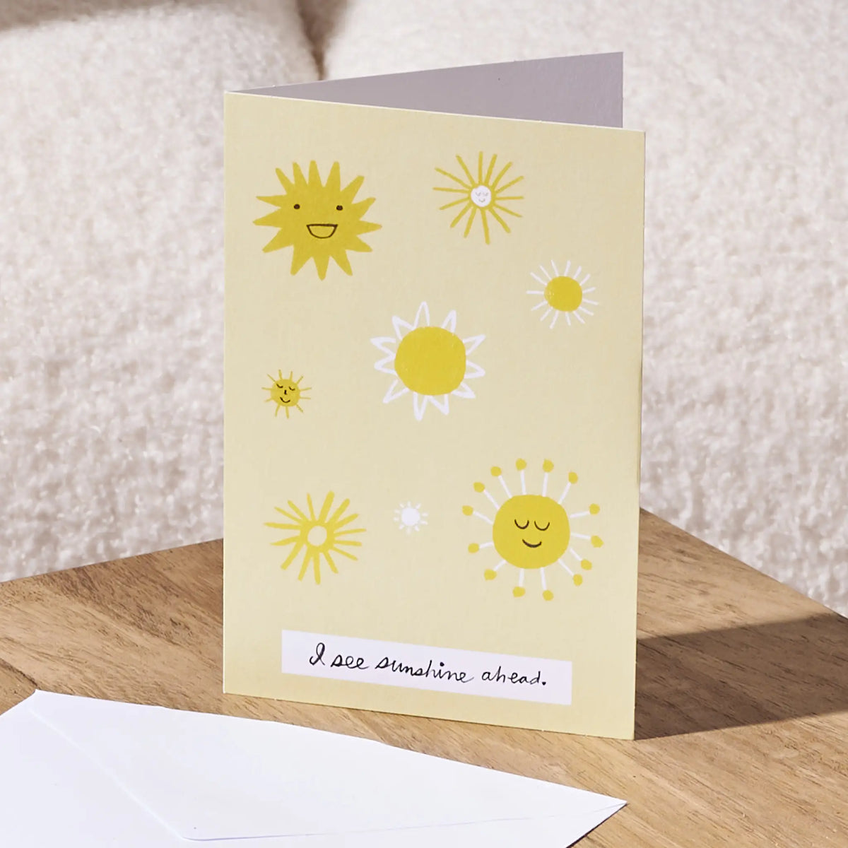 A greeting card with a pale yellow background and playful sun illustrations, standing on a wooden surface with an envelope in front. The card reads, 'I see sunshine ahead.