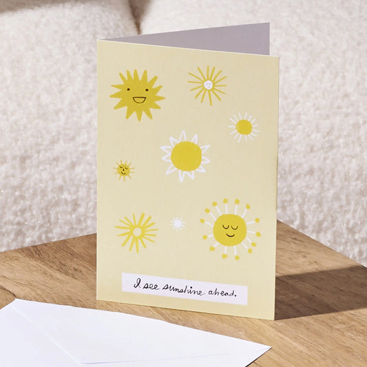 A greeting card with a pale yellow background and playful sun illustrations, standing on a wooden surface with an envelope in front. The card reads, 'I see sunshine ahead.