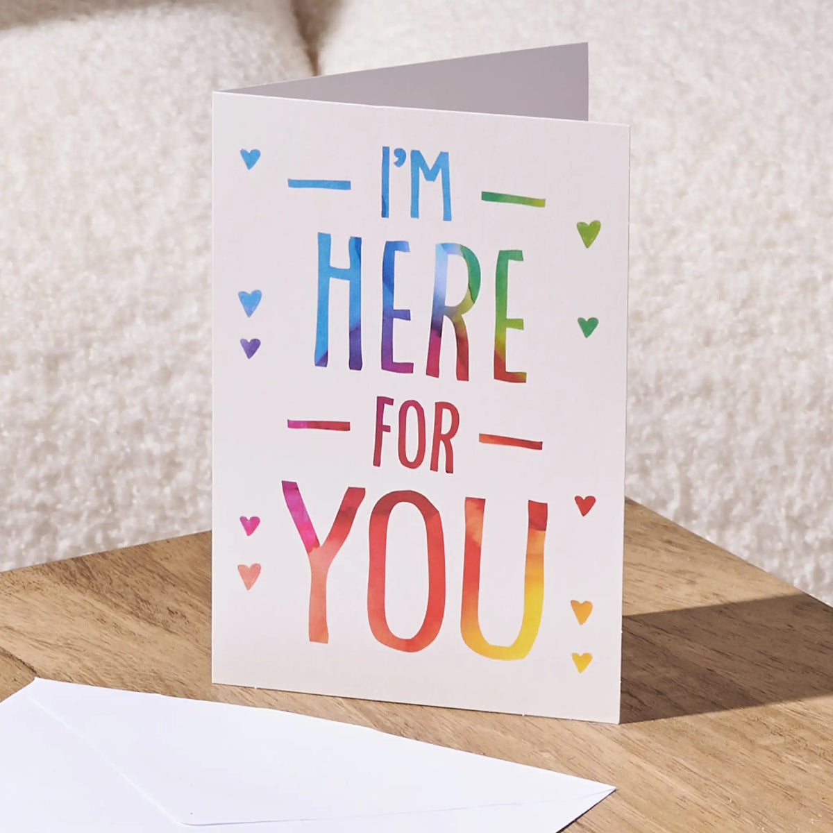 A white greeting card standing open on a wooden surface, printed with the phrase “I'M HERE FOR YOU” in rainbow-colored lettering and small heart accents. A white envelope lies in front, with a soft neutral background behind it.