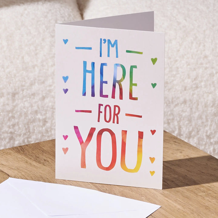 A white greeting card standing open on a wooden surface, printed with the phrase “I'M HERE FOR YOU” in rainbow-colored lettering and small heart accents. A white envelope lies in front, with a soft neutral background behind it.