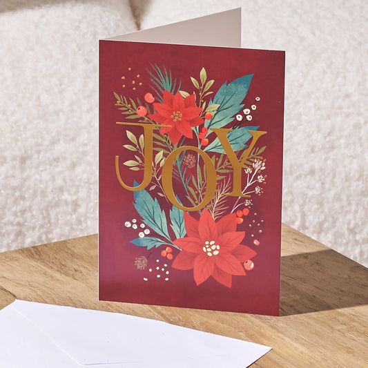 A red holiday greeting card standing upright on a wooden surface, featuring the word “JOY” in large gold letters surrounded by red poinsettias, teal leaves, and small white berries. A white envelope lies in front, with a soft neutral background behind.