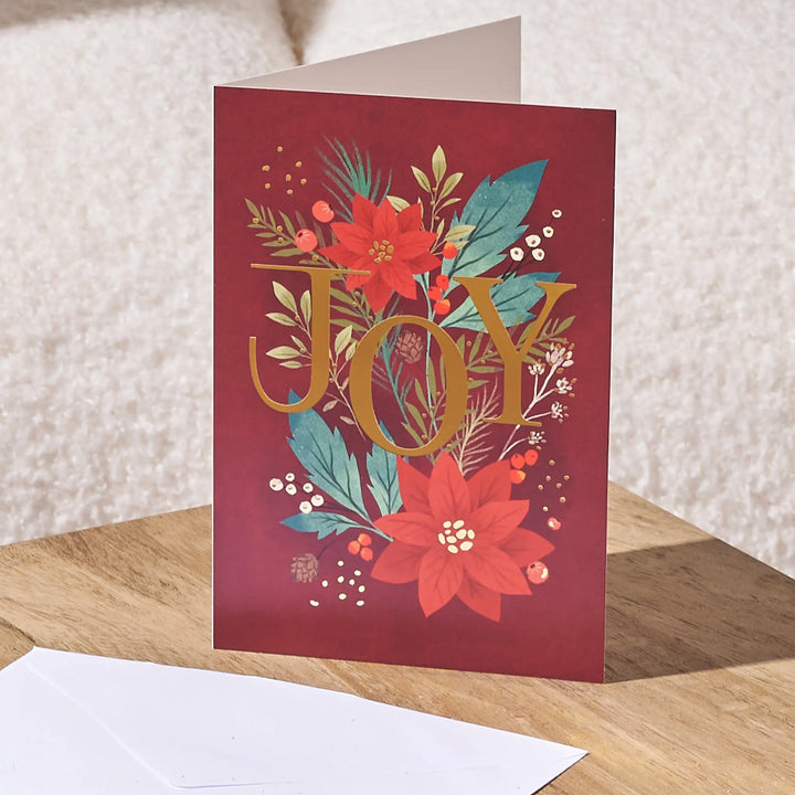 A red holiday greeting card standing upright on a wooden surface, featuring the word “JOY” in large gold letters surrounded by red poinsettias, teal leaves, and small white berries. A white envelope lies in front, with a soft neutral background behind.