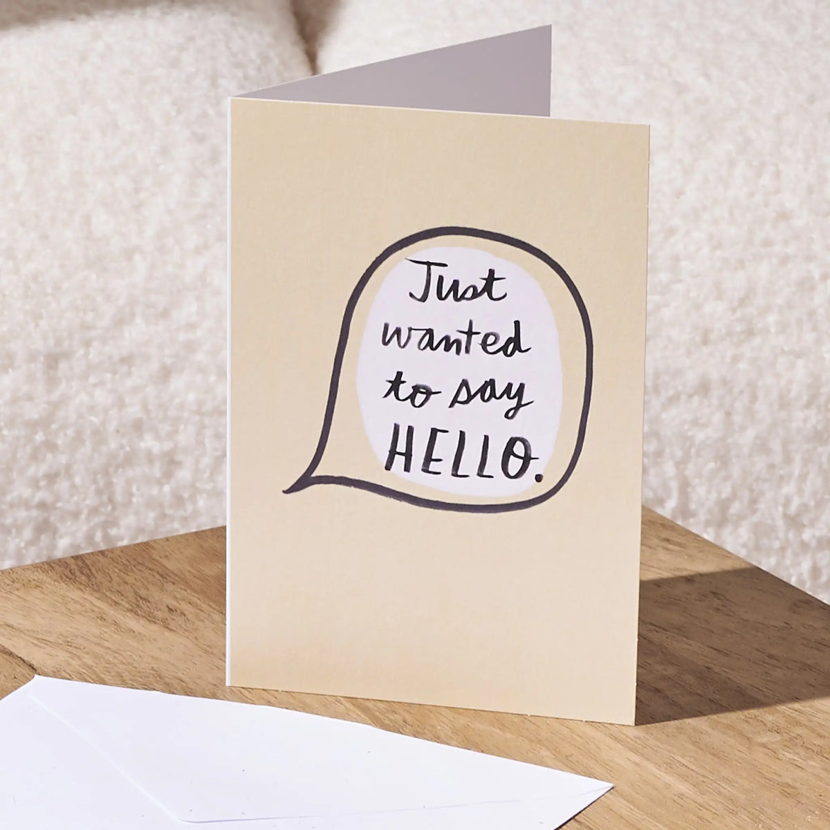 A folded greeting card standing on a wooden surface, with a white envelope in front. The card has a warm cream background and a speech-bubble style graphic with the handwritten phrase 'Just wanted to say HELLO.