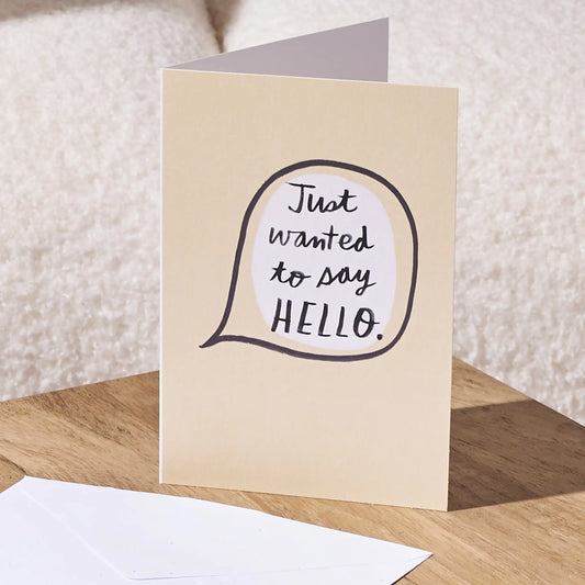 A folded greeting card standing on a wooden surface, with a white envelope in front. The card has a warm cream background and a speech-bubble style graphic with the handwritten phrase 'Just wanted to say HELLO.