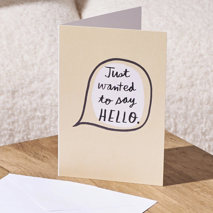 A folded greeting card standing on a wooden surface, with a white envelope in front. The card has a warm cream background and a speech-bubble style graphic with the handwritten phrase 'Just wanted to say HELLO.