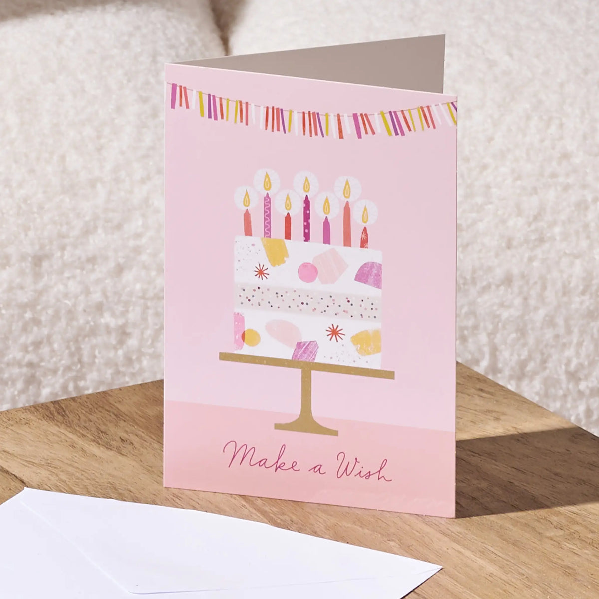 A pink birthday greeting card standing on a wooden surface, featuring an illustrated two-tier cake with candles, bunting, and the words 'Make a Wish'. A white envelope is placed in front of the card, with a soft neutral background.