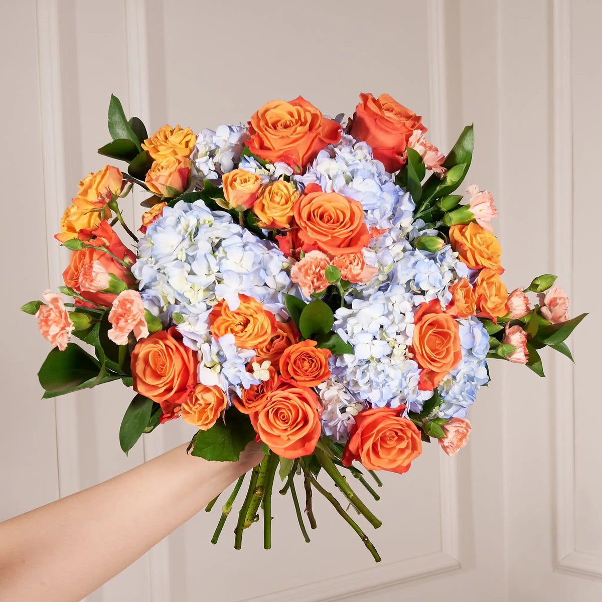 A vibrant bouquet of artificial flowers held by a person's hand against a light beige paneled wall. The bouquet features a balanced mix of bright orange roses, light blue hydrangeas, soft pink carnations, and green foliage.