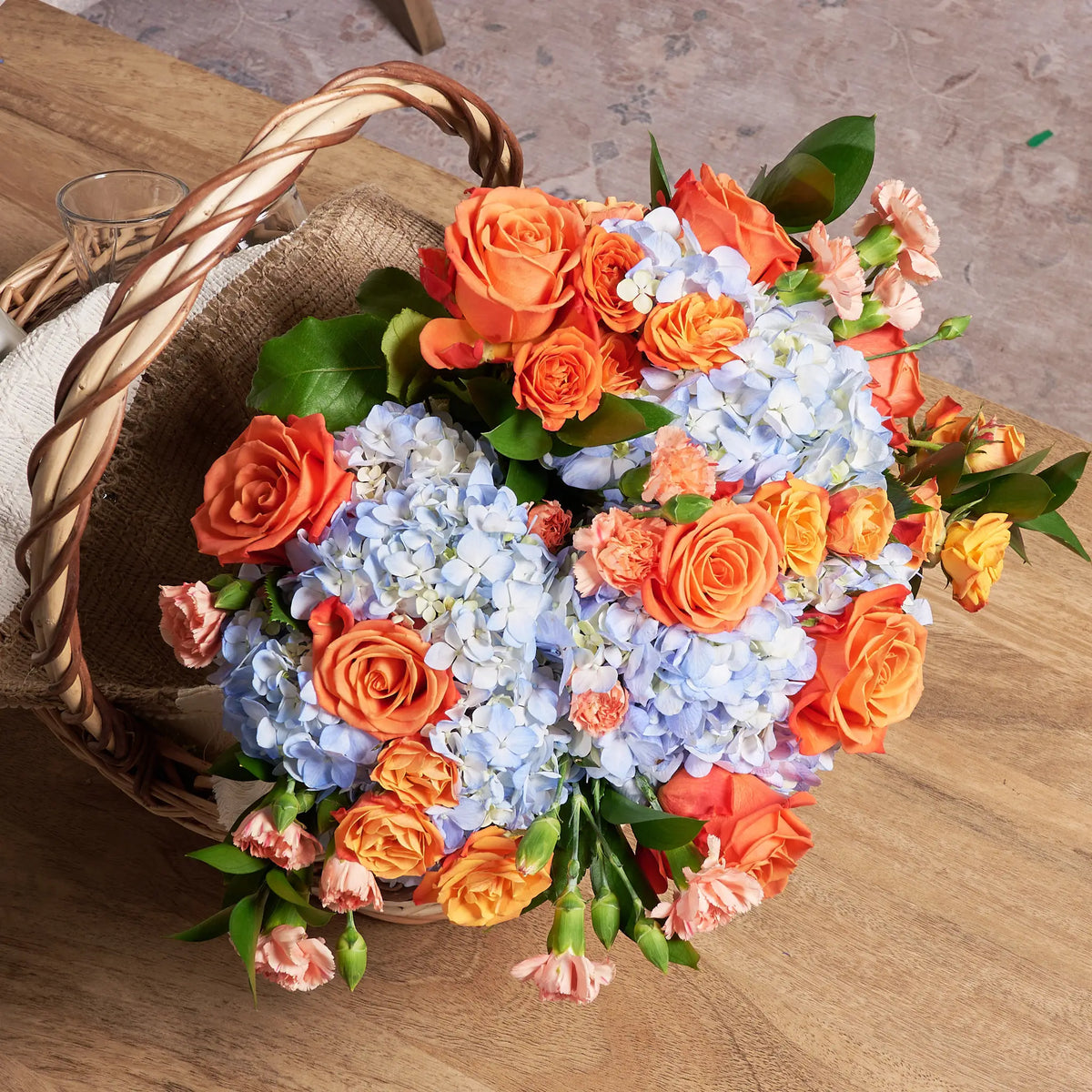 A wicker basket filled with a floral arrangement of light blue hydrangeas, orange roses, pink carnations, and green foliage, set on a wooden table.