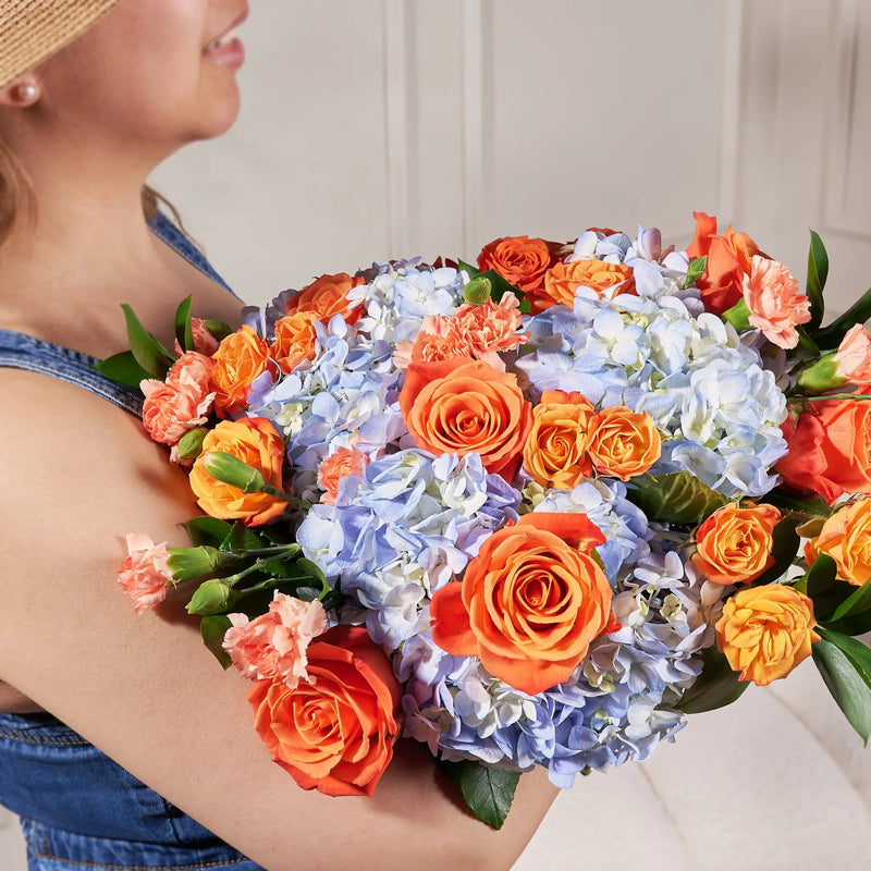A close-up of a woman holding a large bouquet of orange roses, light blue hydrangeas, and pink flowers. The woman is wearing a straw hat and a denim top, and the background is a soft beige indoor setting.