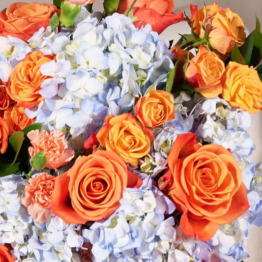A close-up arrangement of vibrant orange roses mixed with light blue hydrangeas and small pink carnations, filling the frame.