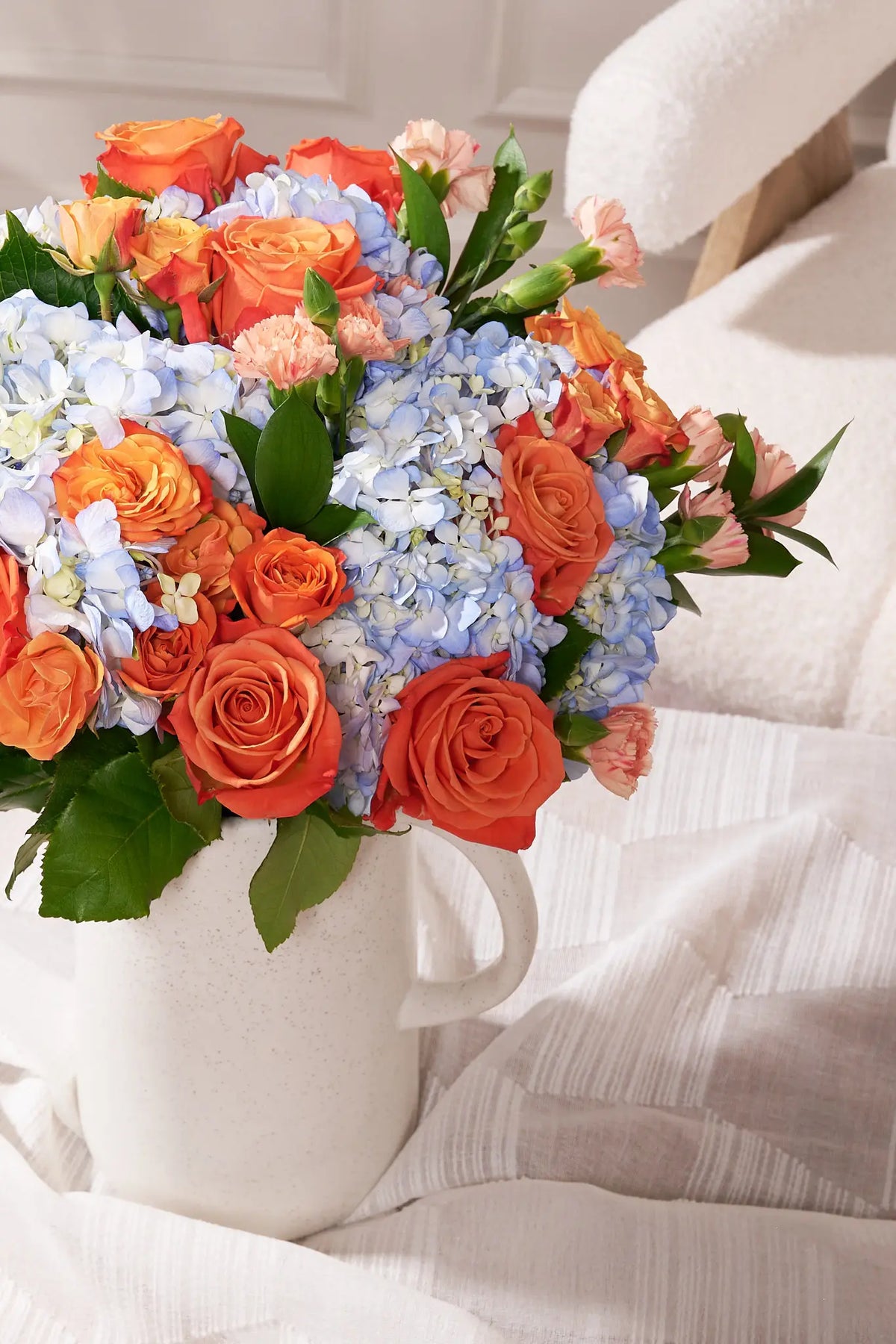 A vibrant floral arrangement featuring orange roses, light blue hydrangeas, and peach carnations with green leaves, presented in a white ceramic pitcher vase on a white textured fabric surface.