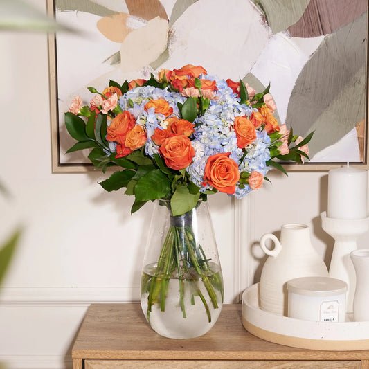 A vibrant artificial flower bouquet featuring orange roses, light blue hydrangeas, and green foliage, arranged in a clear glass vase on a wooden console table next to white ceramic decor.