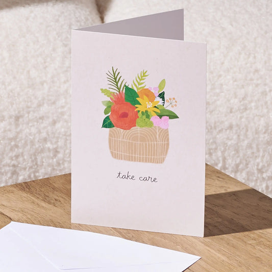 A folded greeting card standing on a wooden surface, printed with a colorful floral basket illustration and the words 'take care' in black script. A white envelope lies in front, with a soft cream textured background behind.