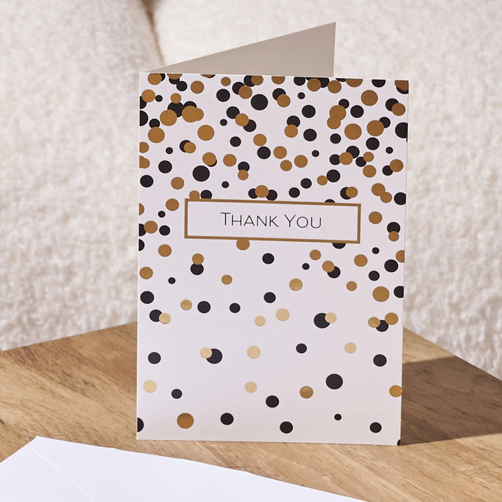 A white thank you card with a scattered gold and black confetti pattern and a central label reading 'THANK YOU', standing on a wooden surface with a white envelope partly visible in front.