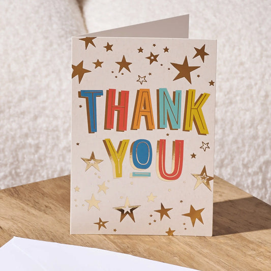 A folded thank you card standing upright on a wooden surface, with a white envelope partly visible in the foreground. The card has a warm cream background decorated with gold stars and the words “THANK YOU” in large, colorful block letters.