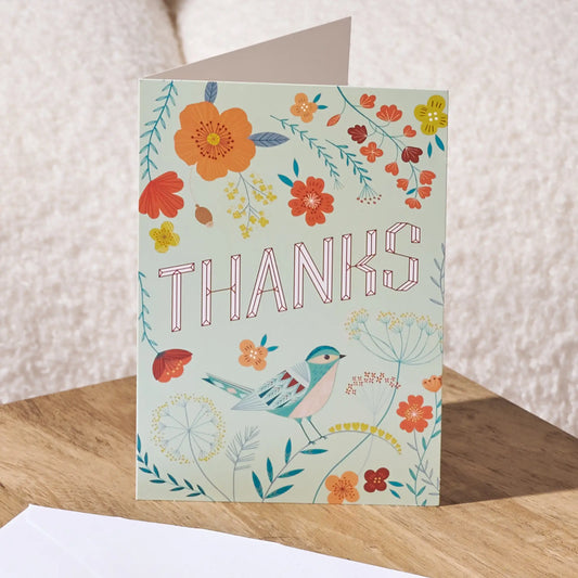 A floral 'THANKS' greeting card with a bird illustration, standing on a wooden surface against a textured cream background.