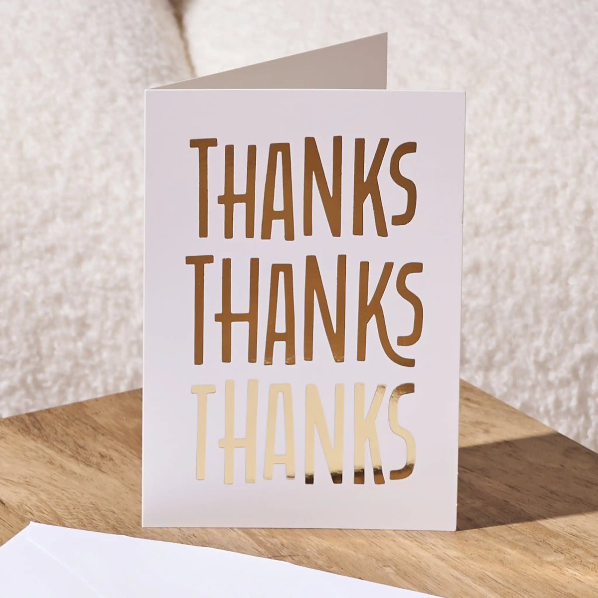 A folded white greeting card standing upright on a wooden surface, with the word 'THANKS' repeated three times in large metallic gold letters. A white envelope is partially visible in the foreground, and a soft beige textured fabric background appears behind it.