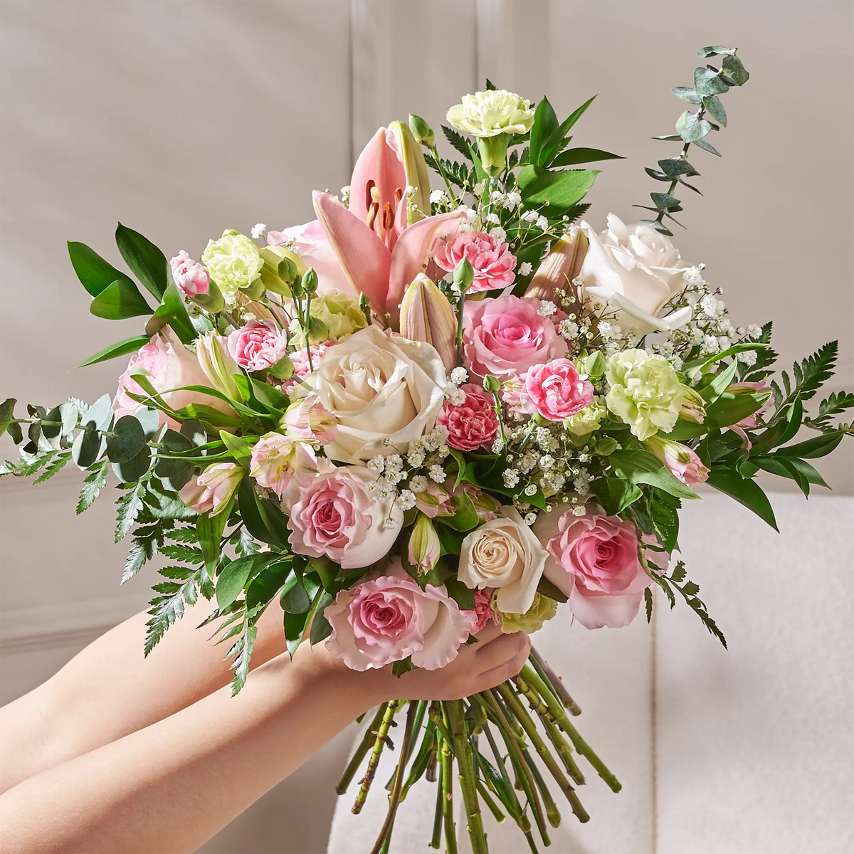 Handheld bouquet of pink and white roses by Gilded Blooms, lilies, and mixed greenery, arranged in a lush, garden-style composition against a soft neutral background.